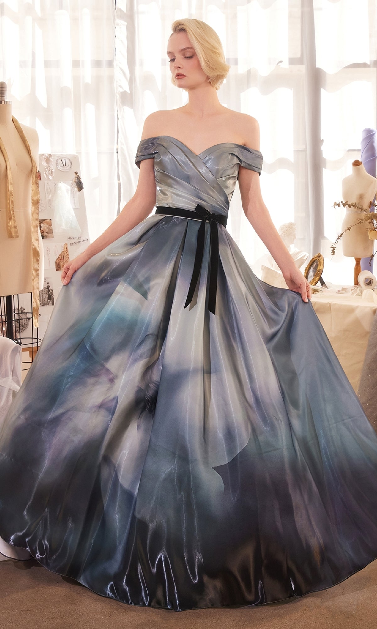 A woman poses in a studio wearing the Andrea & Leo A1431 Off-Shoulder Blue Prom Gown, a floor-length dress with blue and gray florals and a black ribbon belt. Mannequins and sewing materials appear in the background.
